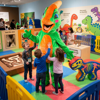 Dinoz le Parasaurolophus mascotte verte et orange, interagissant avec des enfants dans une aire de jeux colorée sur le thème des dinosaures.