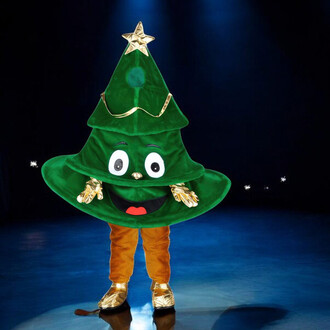 Mascotte sapin cartoon en costume vert vif avec une étoile dorée au sommet, un visage souriant avec des yeux ronds et un sourire, dans un décor sombre avec des lumières projetées. Le personnage porte des gants dorés et une base de costume de couleur marro