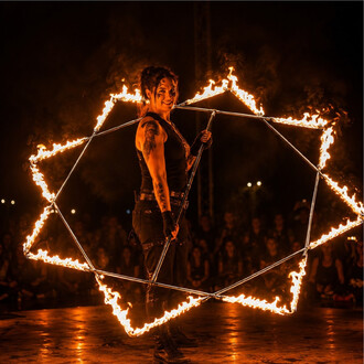 Feuerkünstlerin steht in einem geometrischen Rahmen aus brennendem Metall, nächtliche Performance mit Publikum im Hintergrund.