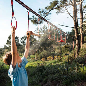 Un enfant s'agrippe à une barre de traction sur un parcours d'obstacles de slackline, entouré d'une nature verdoyante. Des éléments rouges et en bois sont visibles, suspendus à une ligne tendue entre des arbres. Le soleil brille, créant une ambiance dynam