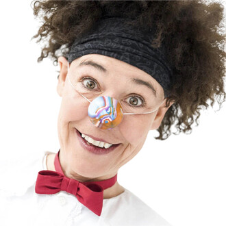 Visage souriant d'une personne portant un nez en silicone coloré, avec des motifs multicolores. Elle a les cheveux frisés, un bandeau noir, et une chemise blanche avec un nœud papillon rouge. Fond blanc.