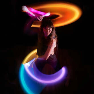 Femme en action avec des nunchakus Lumis Pro LED, les bâtons lumineux en mouvement créent des traînées colorées sur fond sombre, mettant en valeur la dynamique et l'énergie du jonglage.