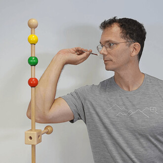 Homme en t-shirt gris, utilisant un instrument pour jongler avec des anneaux colorés (jaune, vert et rouge) fixés sur un support en bois, sur fond clair.