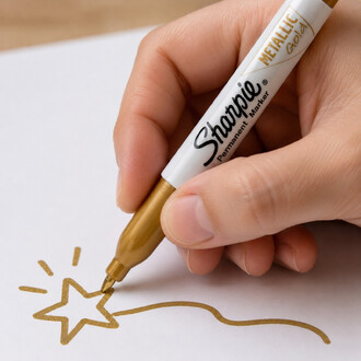 Plan serré sur une main tenant un marqueur Sharpie Metallic Gold en train de dessiner une étoile dorée sur une feuille blanche.