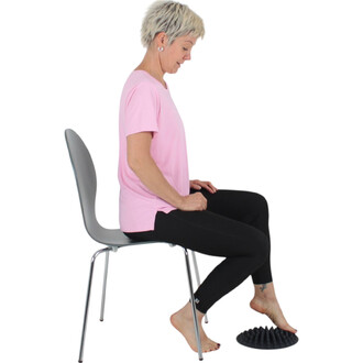 Femme assise sur une chaise, portant un t-shirt rose et un legging noir, avec des pieds nus reposant sur un accessoire de massage en forme de cône. Elle semble concentrée sur son activité. Le fond est blanc, mettant en valeur la posture et l'équipement.