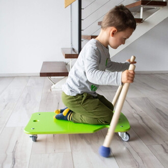 Enfant assis sur un plateau vert à roulettes, tenant des bâtons en bois, dans un intérieur lumineux avec un escalier en arrière-plan.