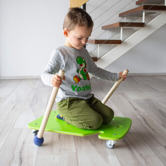 Enfant assis sur une planche verte avec des roulettes, tenant deux bâtons en bois. Il porte un t-shirt à motif de personnage de jeu vidéo et des pantalons verts. La scène se déroule à l'intérieur, avec un escalier visible en arrière-plan.