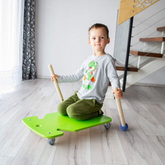 Un enfant assis sur un jouet roulant vert en forme de tortue, tenant deux bâtons en bois. Il porte un t-shirt gris avec un personnage de jeu vidéo et des pantalons verts. L'arrière-plan montre un intérieur moderne avec des escaliers et des rideaux clairs.