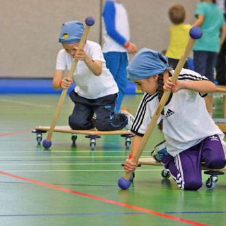 Enfants jouant sur des planches à roulettes dans une salle de sport. Deux enfants sont à genoux, utilisant des bâtons pour propulser leurs planches. Ils portent des t-shirts blancs et des pantalons de sport, avec des foulards bleus sur la tête. L'environn