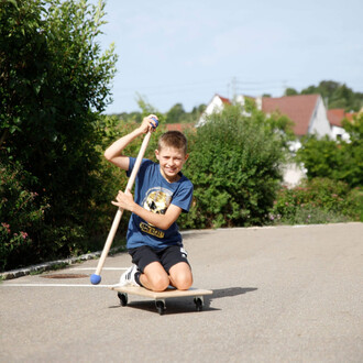 Un garçon souriant est assis sur une planche à roulettes, tenant un bâton avec une boule au bout. Il semble s'amuser en glissant sur une route, entouré de verdure et de maisons en arrière-plan.