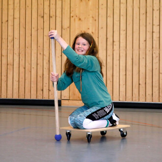 Fille souriante assise sur une planche à roulettes, tenant un bâton avec une boule à son extrémité, dans une salle de sport avec des murs en bois.