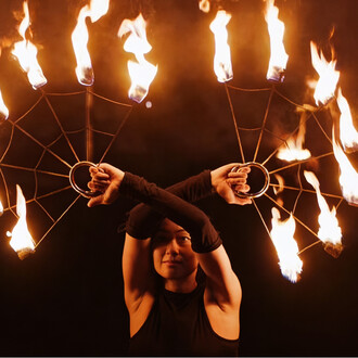 Dois leques de fogo em forma de teia de aranha, segurados por uma pessoa, com chamas emanando das extremidades contra um fundo preto.