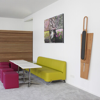 Espace intérieur élégant avec un canapé vert lime, des tables blanches et des poufs roses. Mur décoré avec des tableaux colorés et un élément mural en bois. Sol en moquette grise.