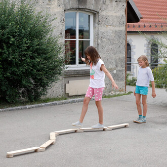 Deux enfants jouent à l'extérieur, l'un d'eux traverse un chemin en bois composé de sections courbes, tandis que l'autre observe. Ils portent des vêtements d'été, avec un mur en pierre et des fenêtres en arrière-plan.