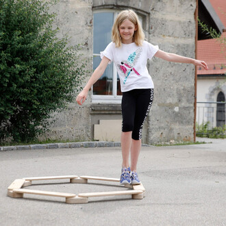 Fille souriante marchant sur un matériel de jonglerie en bois en forme d'hexagone, dans un environnement extérieur. Elle porte un t-shirt blanc avec un motif coloré et un pantalon noir. Arrière-plan d'un mur en pierre et d'une fenêtre.