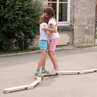 Deux enfants s'amusent en se tenant debout sur une série de planches en bois posées au sol, formant un chemin. L'une porte un T-shirt blanc et un short rose à pois, l'autre un T-shirt blanc et un short bleu. Elles se tiennent l'une contre l'autre avec un 