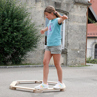 Fille debout sur un cercle en bois, les bras écartés pour maintenir l'équilibre. Elle porte un t-shirt bleu et un short, avec des baskets blanches. En arrière-plan, un mur en pierre et des buissons.