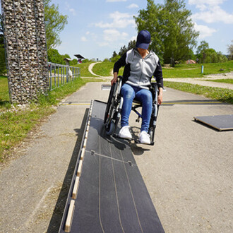 Une personne en fauteuil roulant descend une rampe sur un chemin extérieur, avec un paysage verdoyant et un ciel ensoleillé en arrière-plan. Des éléments de nature et d'infrastructure sont visibles autour d'eux.