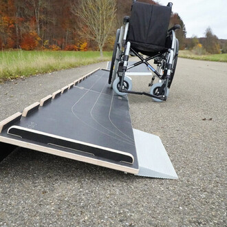 Rampe d'accès pour fauteuil roulant positionnée sur une route avec un fauteuil roulant à proximité. La rampe est construite en matériau composite avec une surface lisse et des rebords, tandis que le fauteuil roulant est noir avec des roues grises. L'envir