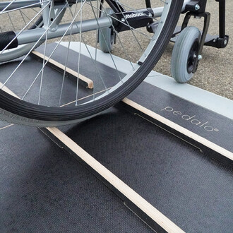 Roues d'un fauteuil roulant sur une surface de jeu en mousse noire, avec des bandes en bois pour guider le mouvement. Un logo pedalo est visible sur le côté du produit.