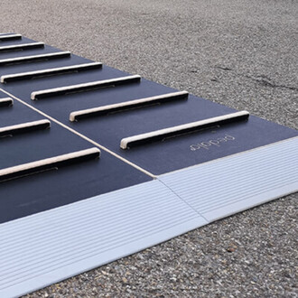 Tapis de jonglerie en noir et gris, avec des bandes en bois surélevées pour la pratique. Le tapis est posé sur une surface rugueuse, avec des détails de texture visibles.