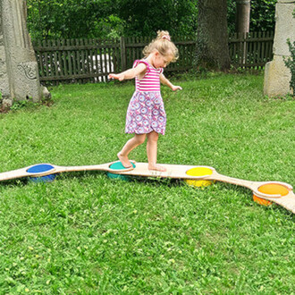 Enfant en bas âge marchant sur une planche d'équilibre colorée, avec des éléments cerclés de différentes couleurs, dans un jardin verdoyant. L'enfant porte une robe à rayures et fleuris et semble concentré tout en maintenant son équilibre.