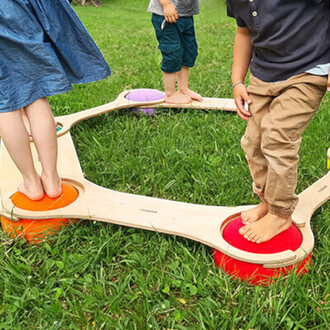 Jeunes enfants jouent pieds nus sur un équipement de jeux en bois, composé de plusieurs plateformes colorées sur une surface herbeuse. L'équipement a une forme circulaire et est conçu pour l'équilibre et l'amusement.