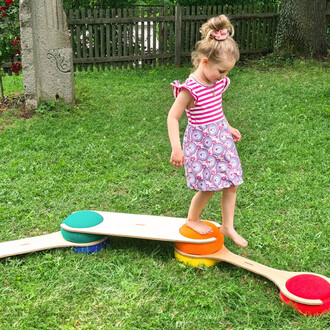 Une petite fille marchant sur un parcours d'équilibre composé de dalles colorées en bois et en mousse, sur une pelouse verte. Elle porte une robe rayée rose et blanche avec un motif floral. Le décor est un jardin avec une clôture en arrière-plan.