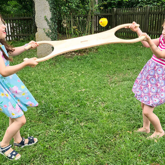 Deux enfants jouent dans un jardin verdoyant, tenant un grand accessoire en bois avec deux trous au centre. Un ballon jaune est en l'air, indiquant qu'ils sont en train de jouer à un jeu amusant. Les enfants portent des vêtements colorés et affichent des 