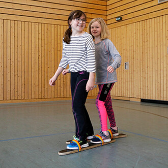 Deux filles se tiennent debout sur une planche de jonglerie, souriantes et équilibrées. L'une porte un t-shirt rayé et l'autre un pull gris, tandis qu'elles sont habillées de pantalons de sport colorés. L'environnement est une salle avec des murs en bois,