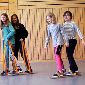 Quatre enfants en mouvement, utilisant des planches de coordination avec des cordes colorées, dans une salle avec des murs en bois. Ils portent des vêtements de sport et semblent s'amuser.