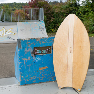 Planche de surf en bois au premier plan, posée près d'un module de skate marqué en bleu avec l'inscription CONCRETE. En arrière-plan, une rampe de skate visible, entourée de verdure et de graffitis colorés.