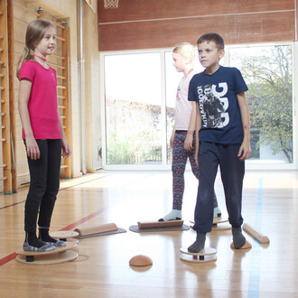 Trois enfants jouent dans une salle de sport. Un garçon en t-shirt bleu et un garçon en t-shirt rose se tiennent debout sur des plateformes de jonglerie. Une fille avec une chemise rose est en train de les regarder. Les enfants sont en chaussettes, et le 