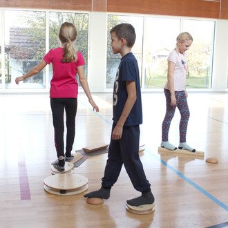 Trois enfants s'exercent sur des équipements de jonglerie ou d'équilibre dans une salle lumineuse. Deux d'entre eux se tiennent sur des plates-formes circulaires, tandis qu'une fille, vêtue d'un t-shirt rose, marche avec précaution. Un garçon portant un t