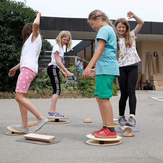 Quatre enfants jouent sur des planches d'équilibre dans un espace extérieur. Ils portent des vêtements colorés et s'amusent, certains souriant tandis qu'ils tentent de maintenir leur équilibre. L'environnement est décontracté, avec des arbres en arrière-p