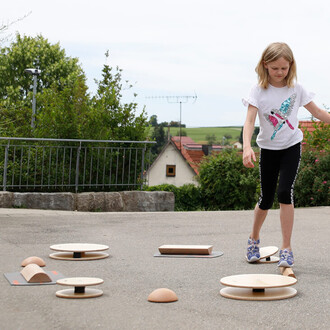 Une fille marche sur un parcours de jonglerie composée de différents équipements en bois, comme des disques et des planches. Elle porte un t-shirt blanc avec un motif coloré et des leggings noirs. L'environnement est extérieur, avec des maisons et des arb