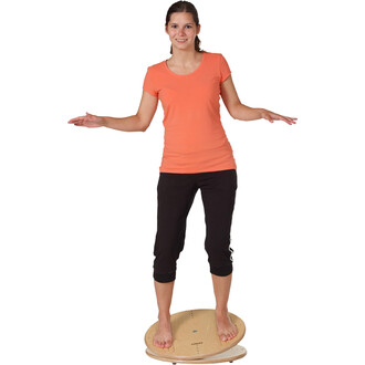 Femme souriante en t-shirt orange et pantalon noir se tenant debout sur une planche d'équilibre en bois, avec les bras écartés pour maintenir son équilibre.