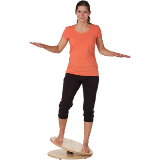 Femme debout sur un wave board en bois, les bras tendus pour maintenir son équilibre. Elle porte un t-shirt orange et un pantalon noir, avec un sourire sur le visage. Le fond est blanc.