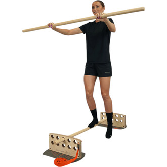 Femme en tenue de sport noire sur un équilibre rigide, tenant une barre en bois au-dessus de sa tête. Elle est pieds nus sur une plateforme en bois avec des supports. L'image met en avant l'utilisation d'un équipement de jonglerie pour améliorer l'équilib