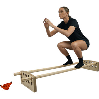 Femme effectuant un squat sur un accessoire en bois de jonglerie, avec les pieds sur une barre horizontale. Elle porte un t-shirt noir et des chaussettes noires, tout en gardant une posture dynamique et concentrée. Un cordon rouge est visible à gauche de 