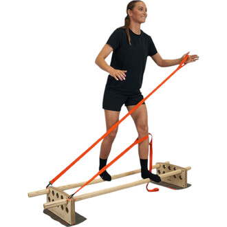 Femme souriante en tenue de sport noirs, en train d'utiliser un équipement de slackline avec des bandes élastiques orange. Elle se tient en équilibre sur une structure en bois, avec une jambe sur la barre et l'autre en l'air, illustrant l'exercice et la p