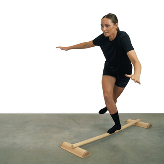 Femme en équilibre sur une barre de jonglage en bois, bras tendus pour maintenir sa posture, vêtue d'un t-shirt noir et de shorts. Le sol est en béton, et la barre est posée sur des supports en bois.