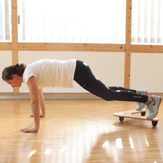 Femme effectuant un exercice de planche sur un plateau de jonglerie, avec les mains au sol et les pieds appuyés sur le plateau, dans un intérieur lumineux. Elle porte un t-shirt blanc et des leggings noirs, avec des chaussures de sport colorées.