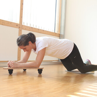 Femme en position de planche sur des genoux, utilisant un plateau à roulettes pour l'exercice, dans une salle lumineuse avec des fenêtres. Elle porte un t-shirt blanc et un legging noir, concentrée sur son entraînement.