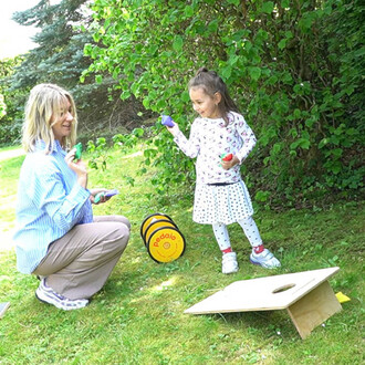 Une femme et une petite fille jouent à l'extérieur dans un jardin. La femme est agenouillée et sourit, tenant des accessoires colorés dans la main. La petite fille, debout, montre une balle rouge et tient une balle bleue, avec un regard joyeux. À côté d'e