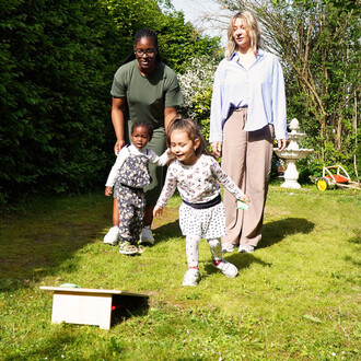 Quatre personnes, dont deux enfants, jouent dans un jardin ensoleillé. Les enfants, vêtus de vêtements colorés, sont en train de courir vers une petite plateforme de jeu en bois, tandis que les adultes les encouragent. En arrière-plan, on peut voir des ar