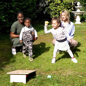 Quatre personnes sont rassemblées dans un jardin ensoleillé. Deux enfants s'amusent au premier plan, l'un portant une salopette à motifs floraux et l'autre une jupe à pois, tandis qu'une fille se prépare à lancer un objet. En arrière-plan, une femme souri