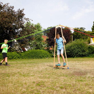 Deux enfants s'amusent dans un parc avec le Pedalo® Course A. L'un d'eux, portant un t-shirt vert, tire sur des cordes colorées, tandis que l'autre, en chemise à carreaux, avance sur la plateforme en bois avec les deux bras agrippés à une structure en boi