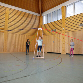 Image de la Pedalo® Course A montrant trois enfants dans une salle de sport. Un enfant au centre utilise l'appareil, tandis que deux autres tiennent des cordes colorées sur les côtés. Le sol est marqué de lignes de jeu et les murs sont en bois, créant une