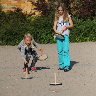 Deux enfants jouant au curling en plein air avec des disques Pedalo. L'un des enfants, une fille aux cheveux longs et blonds, est accroupie et vise un disque au sol, tandis que l'autre, vêtue d'un t-shirt gris et de pantalon turquoise, se tient debout ave